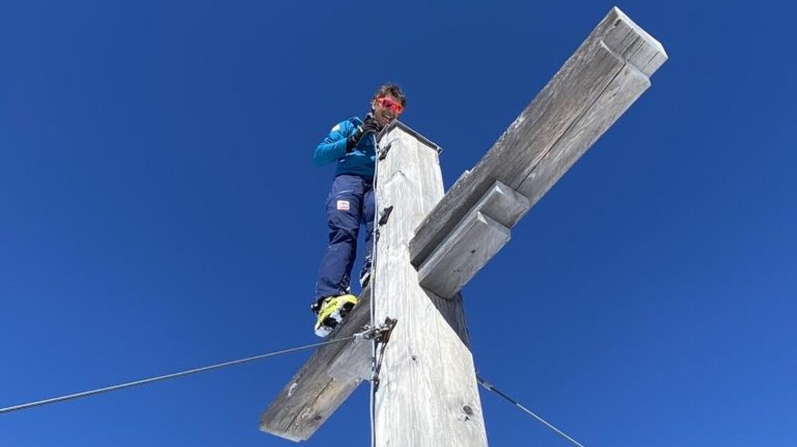 Ein Mensch steht auf einem Holzkreuz in den Bergen. Der Himmel ist klar und blau. | © Peter Perhab