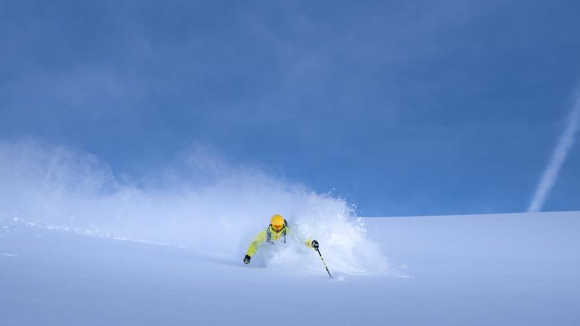 Ein Skifahrer zieht durch unberührten Schnee unter einem klaren blauen Himmel. Der Schnee spritzt um ihn herum und schafft eine dynamische Atmosphäre. | © Peter Perhab