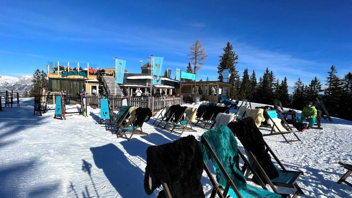 Eine gemütliche Bergwirtschaft im Schnee mit Liegen zum Entspannen. Im Hintergrund erstreckt sich ein klarer blauer Himmel und eine bewaldete Landschaft. | © Knappl's Bergschlössl
