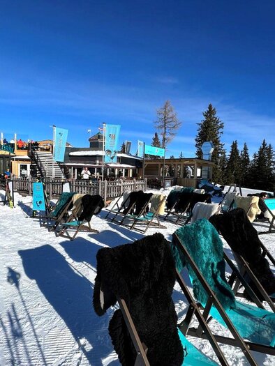 A cozy mountain tavern in the snow with loungers for relaxing. In the background, a clear blue sky and a wooded landscape stretch out. | © Knappl's Bergschlössl