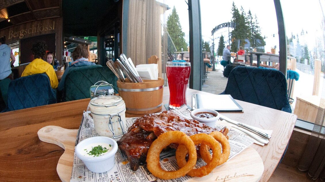 Ein köstlicher Teller mit Rippchen, serviert mit Zwiebelringen und Sauces. Im Hintergrund sieht man eine gemütliche Atmosphäre und eine Aussicht auf die Berge. | © Knappl's Bergschlössl