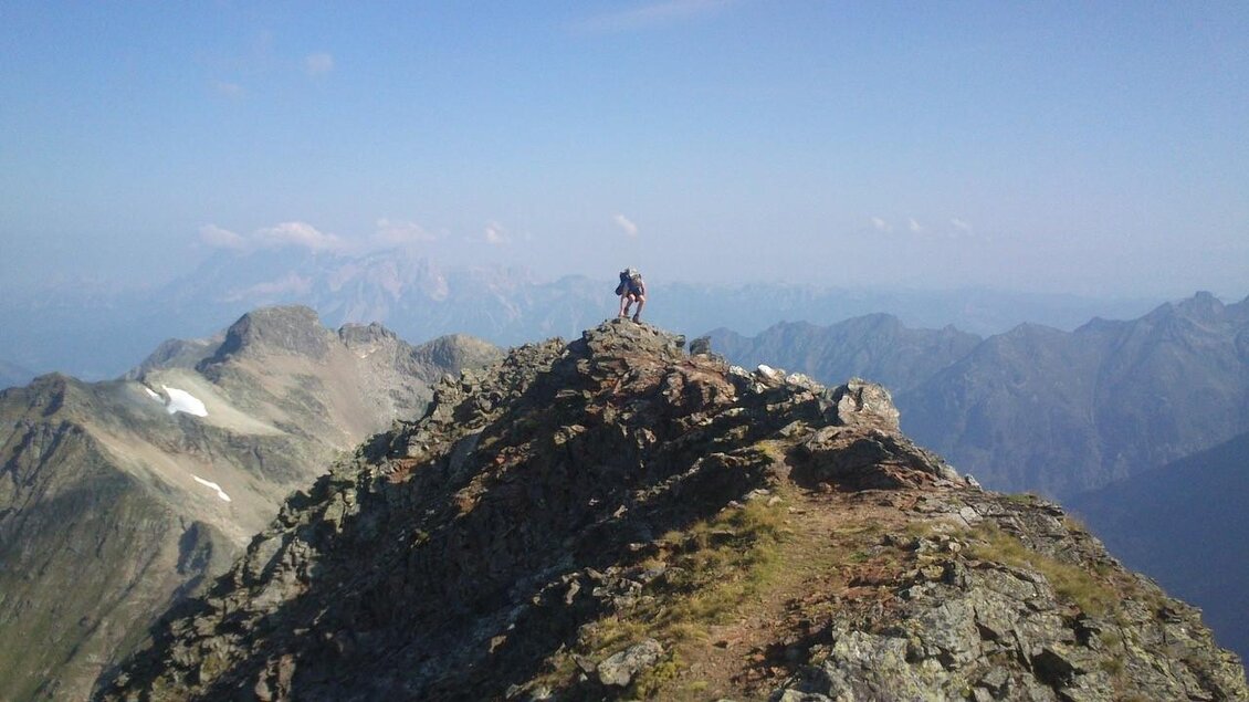 Ein Wanderer steht auf einem schmalen Bergrücken mit beeindruckender Aussicht auf Bergketten. Der Himmel ist klar und die Landschaft ist von grünen und grauen Tönen geprägt. | © Martin Huber