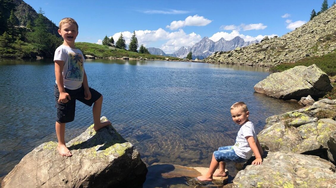 Zwei fröhliche Jungen stehen auf Felsen am Ufer eines klaren Sees. Im Hintergrund sind grüne Hügel und Berge bei schönem Wetter zu sehen. | © Martin Huber