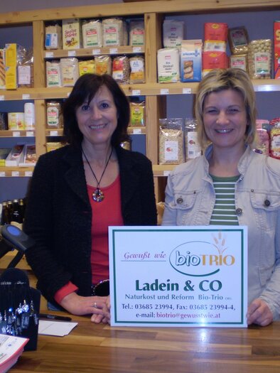 Two women are smiling behind a sales counter in a store. In the background, shelves with various food and products are visible. | © Bio Trio