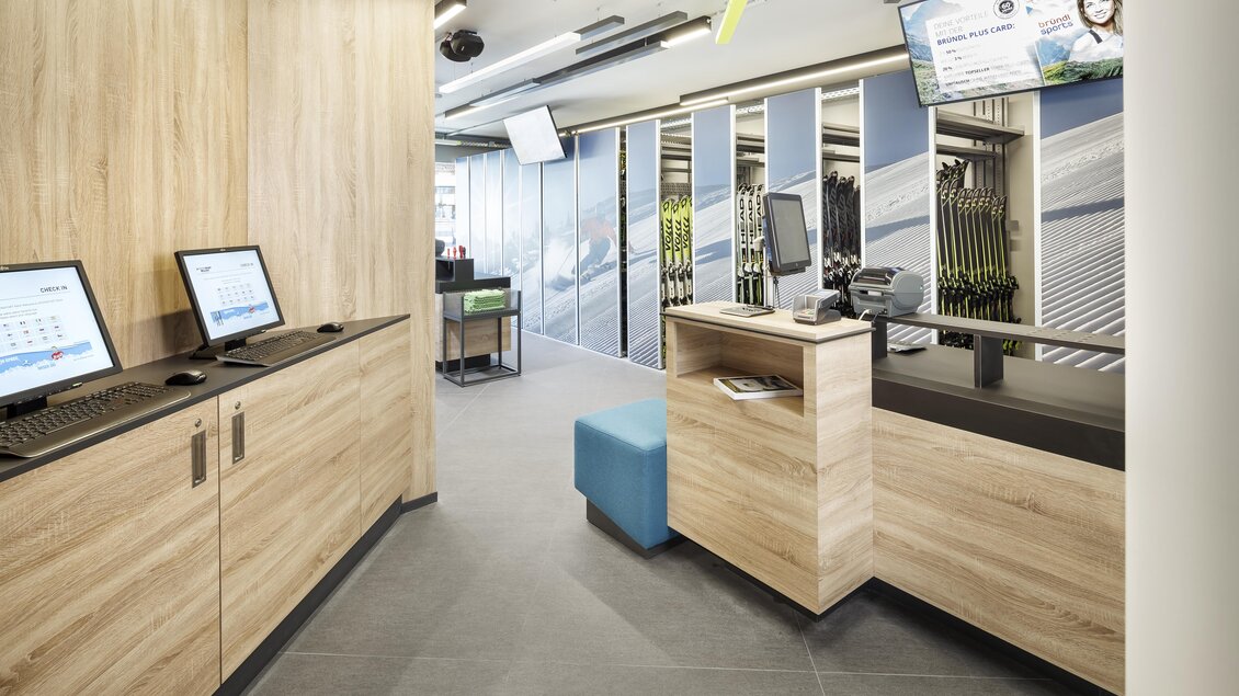 Ein moderner Empfangsbereich mit Holzvertäfelung und digitalen Terminals. Die Atmosphäre ist hell und einladend, mit Grafiken an den Wänden und einem Empfangstresen. | © Bründl Sports / Christian Wöckinger