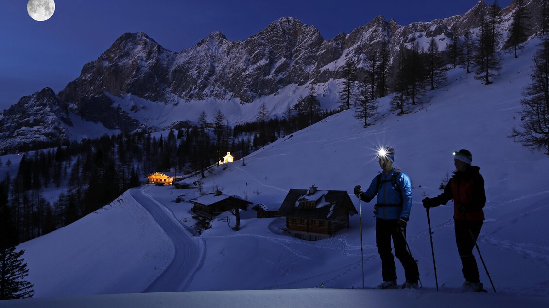 Zwei Skitourengeher wandern nachts durch eine schneebedeckte Landschaft. Der volle Mond beleuchtet die Berge im Hintergrund und ein paar Häuschen im Tal. | © Herbert Raffalt
