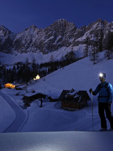 Zwei Skitourengeher wandern nachts durch eine schneebedeckte Landschaft. Der volle Mond beleuchtet die Berge im Hintergrund und ein paar Häuschen im Tal. | © Herbert Raffalt