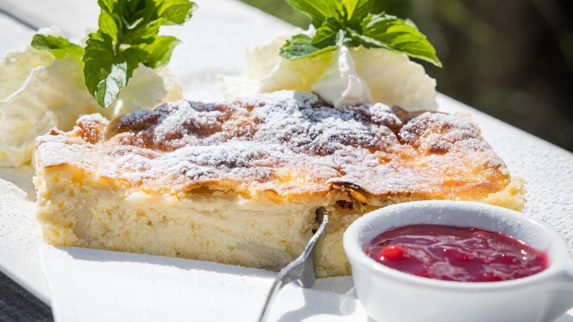 Ein Stück Kuchen mit Puderzucker und frischer Minze, serviert auf einem weißen Teller. Dazu gibt es eine kleine Schale mit roter Marmelade. | © Netzwerk Kulinarik wildbild