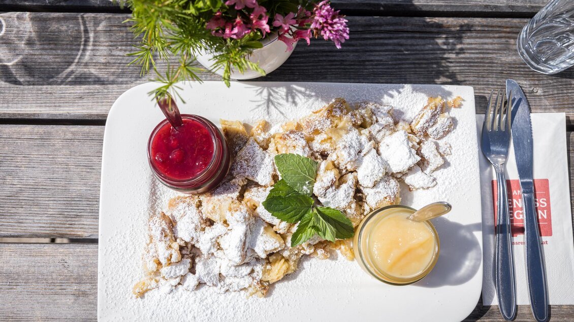 Ein köstlicher Kaiserschmarren auf einem weißen Teller, garniert mit Puderzucker und frischen Kräutern. Daneben befinden sich zwei kleine Gläser mit fruchtigen Soßen. | © Netzwerk Kulinarik wildbild