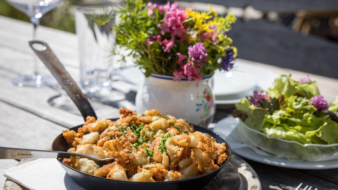 Ein rustikales Gericht in einer Pfanne, umgeben von frischen Kräutern. Neben dem Essen steht ein bunter Blumenstrauß und ein Salatteller. | © Netzwerk Kulinarik wildbild