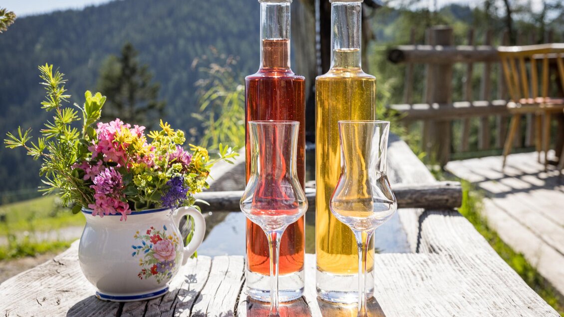 Zwei Flaschen mit verschiedenen Spirituosen stehen auf einem Holztisch. Neben ihnen steht eine Blumenvase mit bunten Blumen vor einer malerischen Berglandschaft. | © Netzwerk Kulinarik wildbild