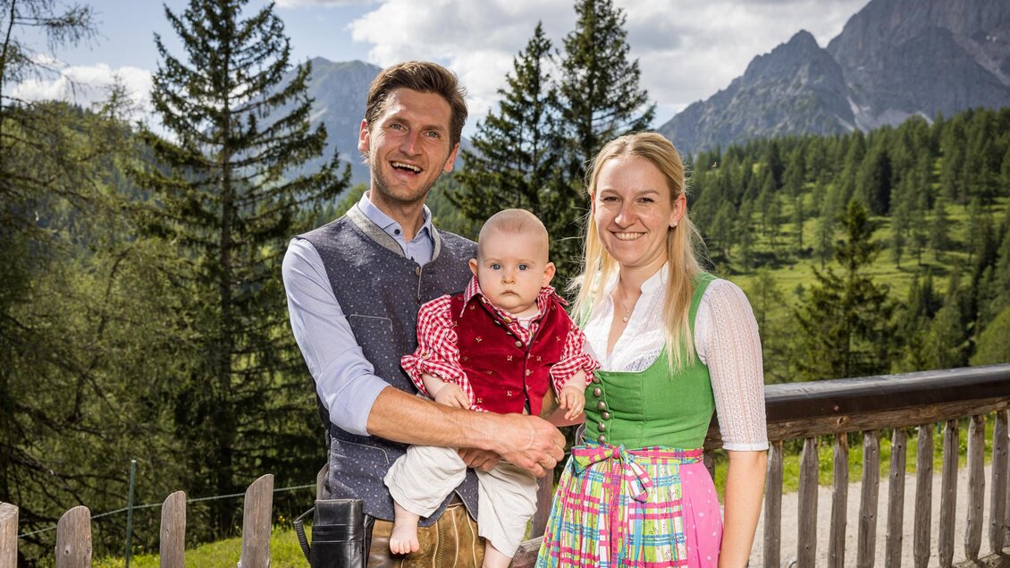 Ein fröhliches Paar steht mit einem Baby vor einer malerischen Berglandschaft. Die Erwachsenen tragen traditionelle Trachten und die Natur ist grün und sonnig. | © Netzwerk Kulinarik wildbild