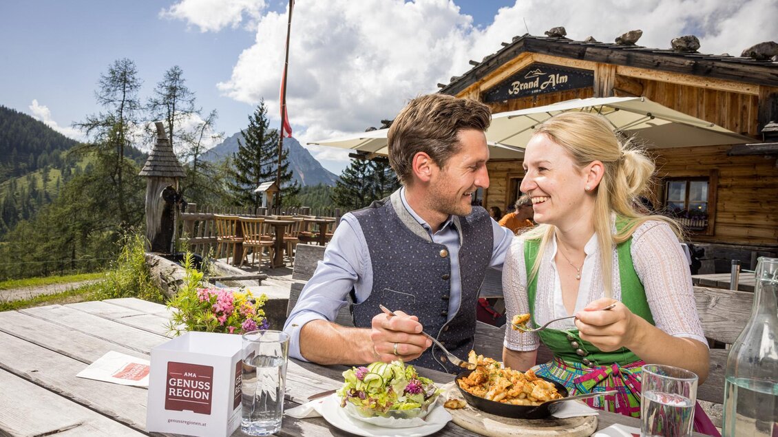Ein paar genießt ein romantisches Essen im Freien auf einer Terrasse. Im Hintergrund sind Berge und eine Hütte zu sehen. | © Netzwerk Kulinarik wildbild