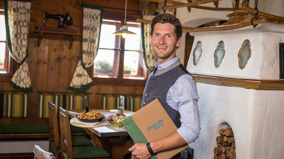 Ein freundlicher Kellner steht in einem gemütlichen Restaurant mit traditionellen Holzmöbeln. Im Hintergrund sind die Tische mit leckerem Essen gedeckt. | © Netzwerk Kulinarik wildbild