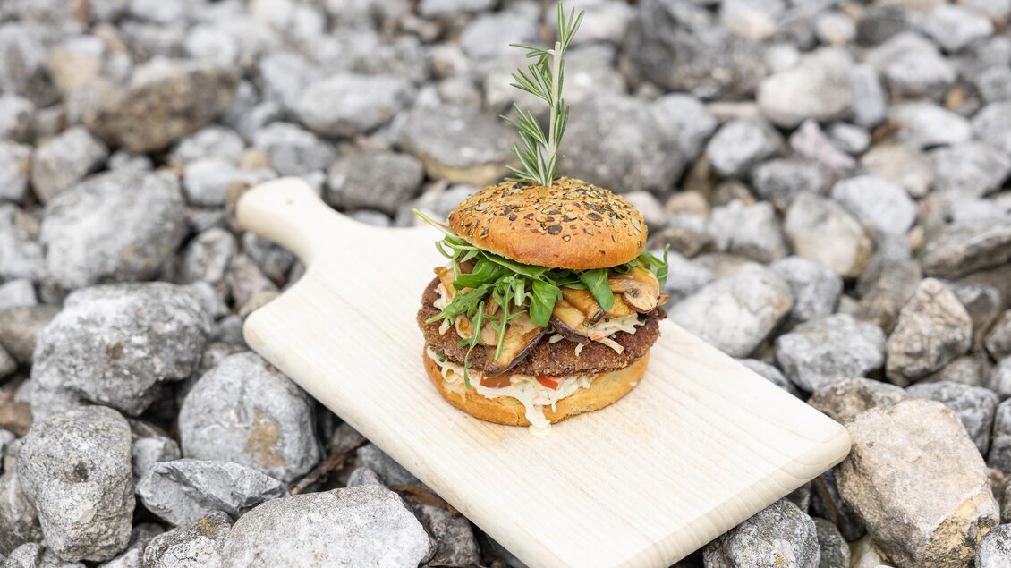 Ein appetitlicher Burger mit frischen Zutaten auf einem Holzbrett. Der Burger ist mit Kräutern dekoriert und liegt auf einem Untergrund aus Kies. | © Martin Huber