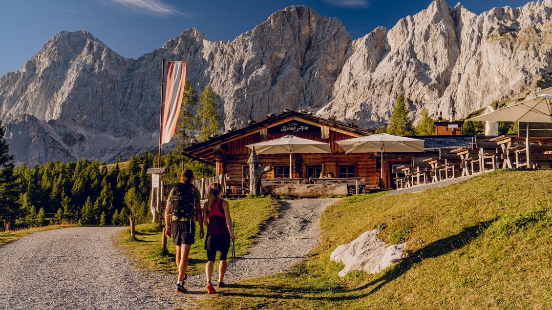 Ein Paar wandert auf einem Schotterweg zu einer Berghütte. Im Hintergrund sind majestätische Berge sichtbar. | © Christine Höflehner