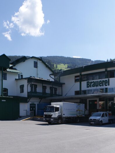 A view of the Schladming brewery with modern buildings and surrounding mountains. The scene also shows a lively atmosphere with several vehicles and people. | © Gerhard Pilz