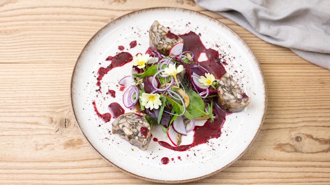 Ein kunstvoll angerichteter Teller mit einer bunten Salatmischung, verziert mit essbaren Blumen und roten Zwiebeln. Dazu gibt es eine dunkle Sauce und mehrere kleine Stücke, die an Fleisch oder Wurst erinnern. | © ©Martin Huber