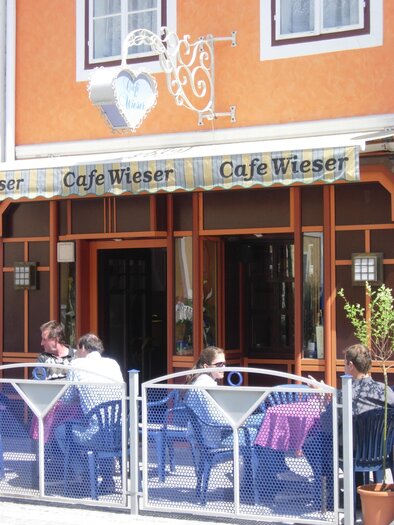 A cozy café with an orange facade. Guests are sitting outside at tables and enjoying the sun. | © Café Wieser
