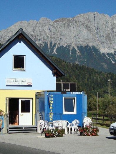 A small, colorful building near a road, surrounded by mountains. In front of the building, there are some chairs and tables outdoors.
