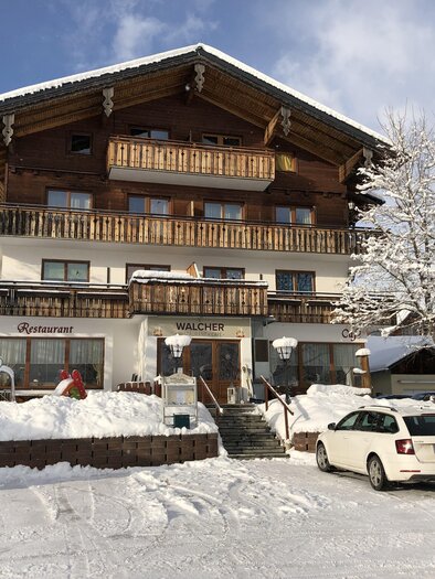 Ein gemütliches Chalet im Winter mit schneebedecktem Grundstück. Parkende Autos stehen vor dem Haus, umgeben von einer winterlichen Landschaft. | © Café Restaurant Walcher