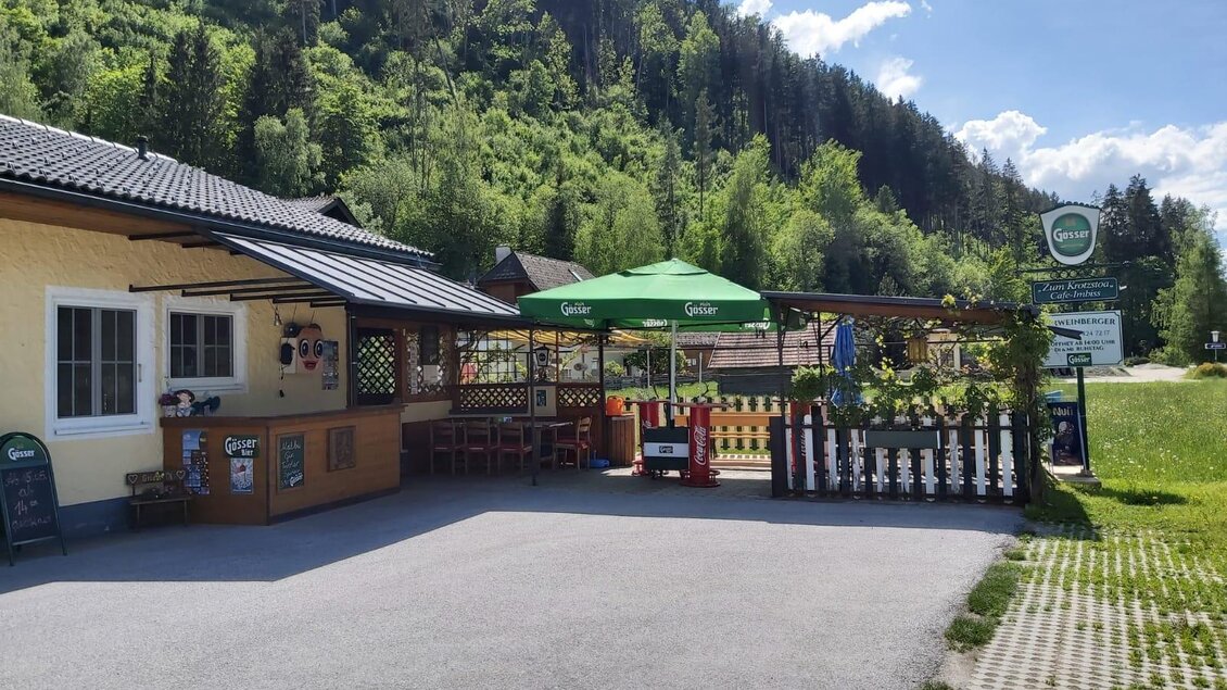 Ein gemütliches Restaurant mit grünen Sonnenschirmen und einer einladenden Terrasse. Im Hintergrund sind bewaldete Hügel und ein blauer Himmel zu sehen. | © Sabine Schweinberger