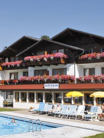 A charming hotel with blooming balconies and a clear swimming pool. The surrounding area is green and inviting, perfect for a relaxing vacation. | © Café-Restaurant Alpenbad