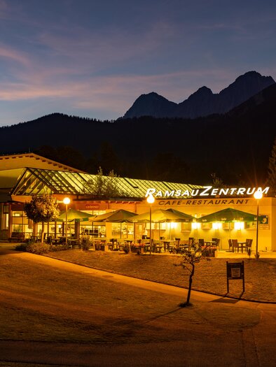 A cozy café-restaurant with a lit outdoor area amidst a picturesque mountain landscape. The surroundings are adorned with trees and gentle hills. | © Hans-Peter Steiner
