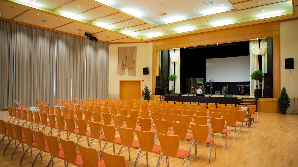 Ein großer Veranstaltungsraum mit vielen Stühlen in Reihen. Auf der Bühne stehen Musikinstrumente und ein Projektor. | © Hans-Peter Steiner