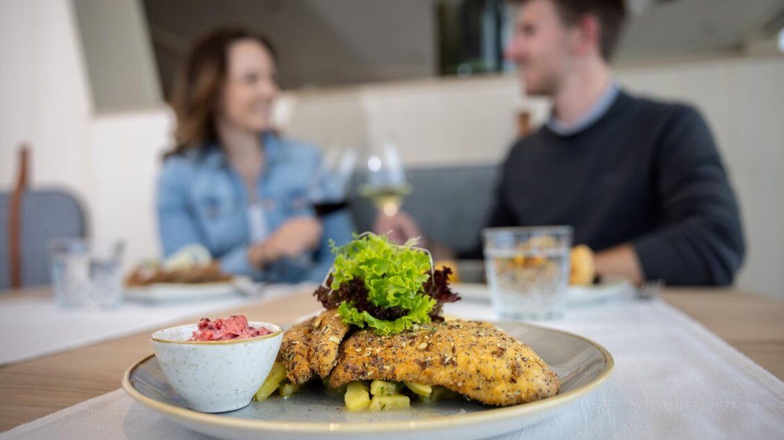 Ein köstlicher Teller mit gegrilltem Hähnchen und Salat auf einem Tisch. Im Hintergrund sind zwei Menschen zu sehen, die einander ansprechen und ein Glas Wein halten. | © Harald Steiner