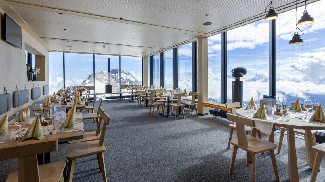 Ein elegantes Restaurant mit großen Fenstern, das einen Blick auf Berge und Wolken bietet. Die Tische sind schön gedeckt und schaffen eine angenehme Atmosphäre. | © Harald Steiner