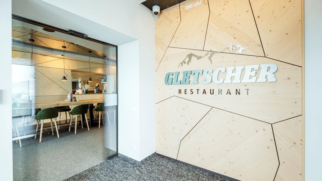 Der Eingang zum Gletscher Restaurant ist modern und einladend gestaltet. Die Wand ist aus Holz mit dem Schriftzug „Gletscher Restaurant“ in dekorativen Buchstaben. | © Christoph Huber