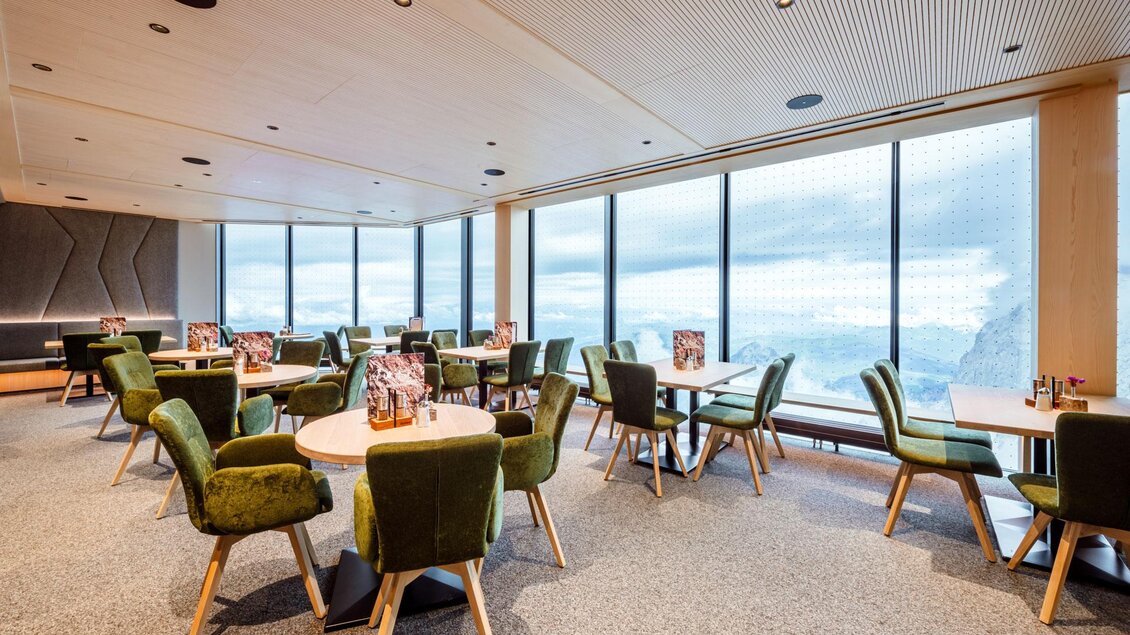 Ein modernes Restaurant mit großen Fenstern und Blick auf die Berge. Die Einrichtung besteht aus runden Tischen und modernen, grünen Stühlen. | © Christoph Huber