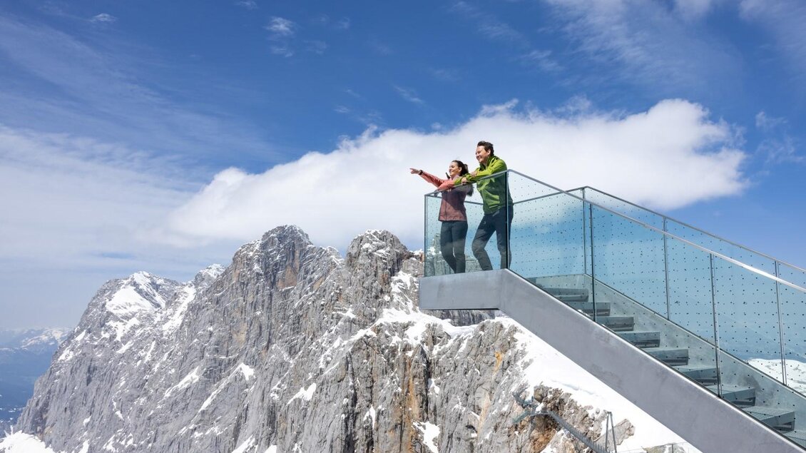 Ein Paar steht auf einer Aussichtsplattform in den Bergen und zeigt auf die Landschaft. Der Himmel ist blau und die Berge sind schneebedeckt. | © Harald Steiner