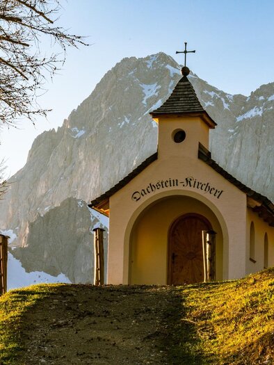 Chapel on a grassy hill, dyed in orange autumn colors. In the background, mountains and blue sky can be seen. | © Christine Höflehner