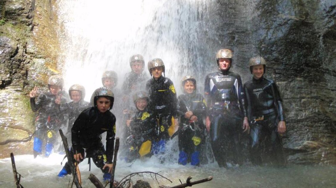Eine Gruppe von Personen steht vor einem Wasserfall. Sie tragen Neoprenanzüge und Helme, während Wasser über sie fließt. | © Martin Seebacher