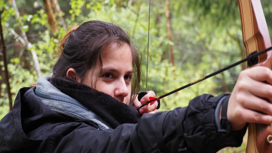 Ein junges Mädchen zielt mit einem Bogen im Wald. Die grünen Bäume im Hintergrund schaffen eine ruhige Atmosphäre. | © Martin Seebacher