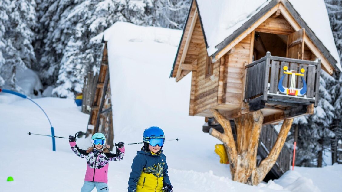 Zwei Kinder fahren Ski auf einer verschneiten Piste. Im Hintergrund steht ein kleine Holzhütte umgeben von hohen Bäumen. | © Ski Galsterberg