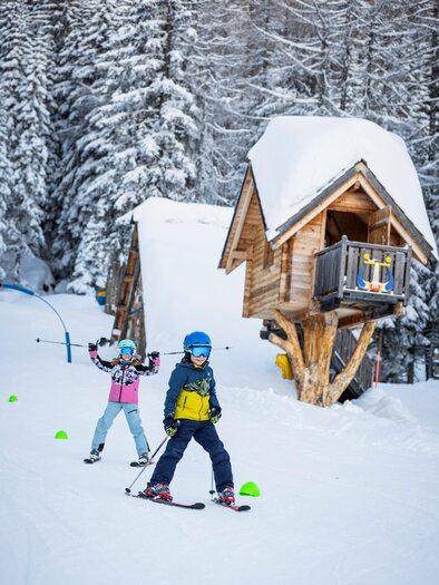 Two children are skiing on a snowy slope. In the background, there is a small wooden cabin surrounded by tall trees. | © Ski Galsterberg