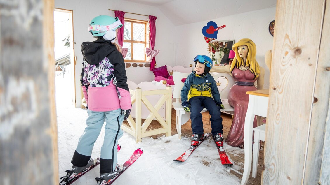 Zwei Kinder in Skiausrüstung stehen in einem gemütlichen Raum mit Schnee. Im Hintergrund sitzt eine große Puppe an einem Tisch. | © Ski Galsterberg
