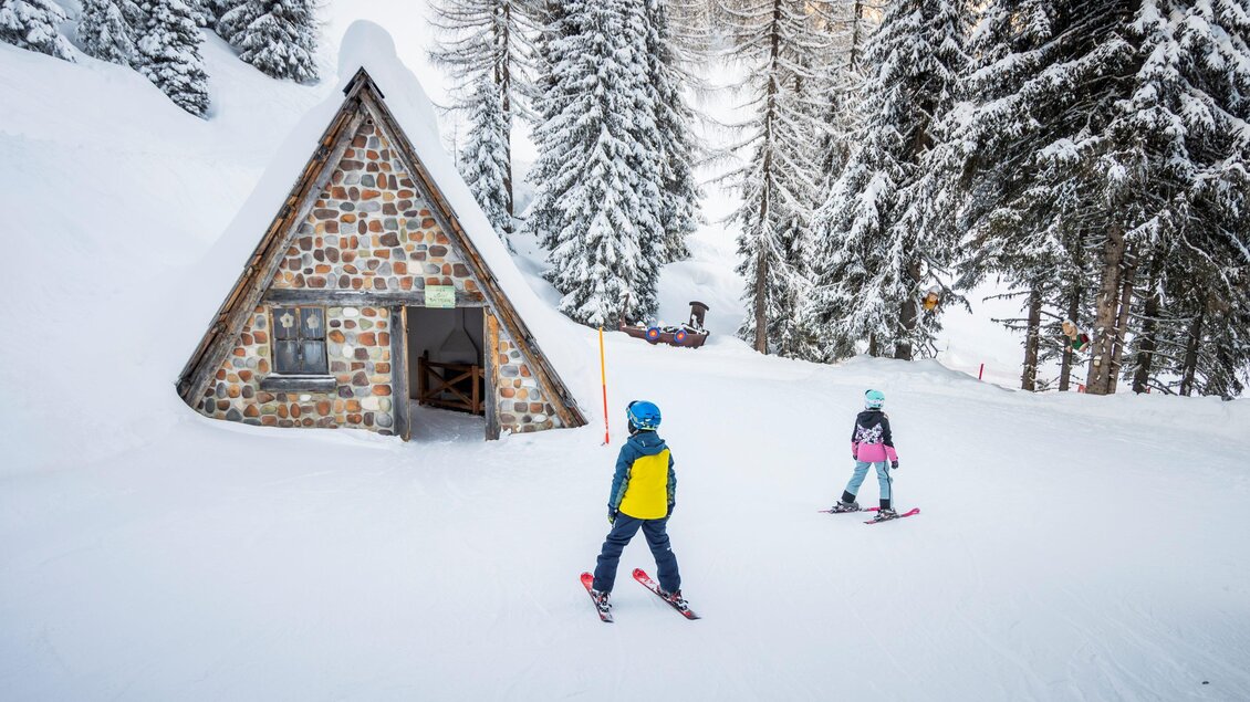 Ein Winterlandschaft mit schneebedeckten Bäumen und einem kleinen, charmanten Holzhaus. Zwei Skifahrer fahren eine sanfte Piste hinunter.