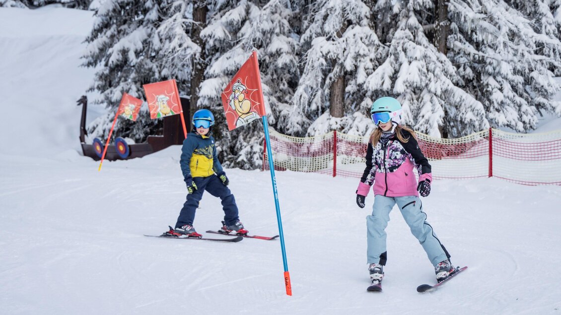 Zwei Kinder fahren Ski im verschneiten Gelände. Im Hintergrund sind einige verschneite Bäume und Fahnen sichtbar. | © Ski Galsterberg