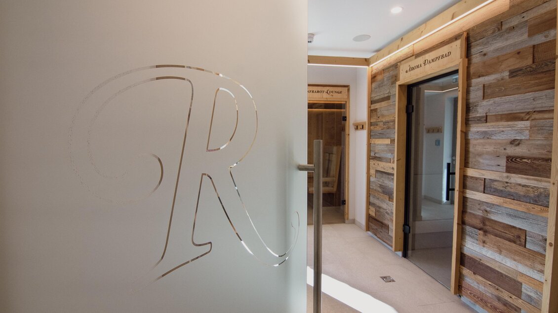 Ein eleganter Innenraum mit einer gläsernen Tür und einem stilisierten „R“. Die Wände sind mit rustikalem Holz verkleidet. | © Rösslhof