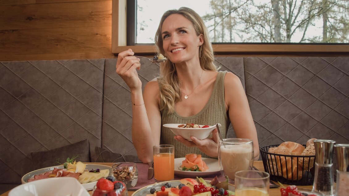 Eine lächelnde Frau sitzt in einem Restaurant und genießt ihr Frühstück. Auf dem Tisch sind verschiedene Speisen und Getränke angerichtet. | © Rösslhof