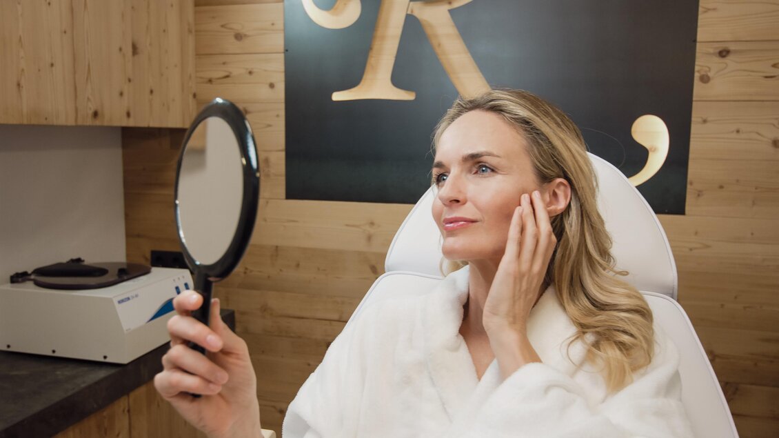 Eine Frau in einem weißen Bademantel betrachtet sich im Spiegel. Sie ist in einem gepflegten Wellness-Bereich. | © Rösslhof