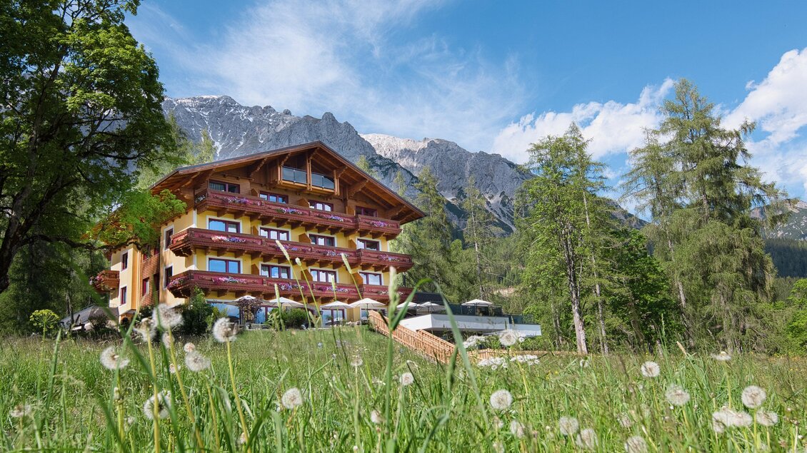 Ein schönes Holzgebäude inmitten von grünen Wiesen und Bäumen. Im Hintergrund sind majestätische Berge unter einem blauen Himmel sichtbar. | © Rösslhof
