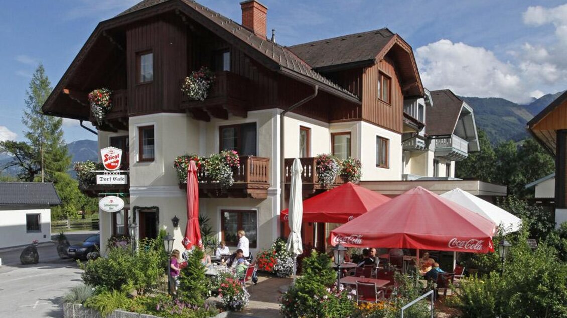 Ein hübsches Gasthaus mit einem einladenden Außenbereich. Es gibt Tische mit roten Sonnenschirmen und Fahrräder, die vor dem Gebäude stehen. | © Dorf Café