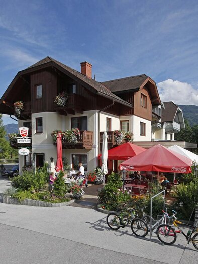 Ein hübsches Gasthaus mit einem einladenden Außenbereich. Es gibt Tische mit roten Sonnenschirmen und Fahrräder, die vor dem Gebäude stehen. | © Dorf Café