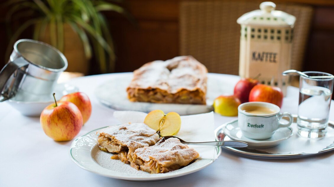 Ein Tisch gedeckt mit frischem Apfelstrudel, Äpfeln und einer Tasse Kaffee. Im Hintergrund steht eine Kaffeemaschine und eine grüne Pflanze. | © Dorf Café