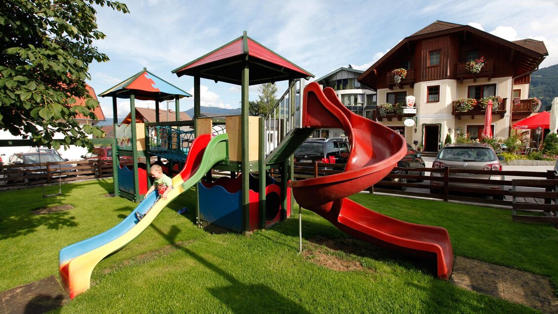 Ein bunter Spielplatz mit Rutschen und Spielgeräten steht auf einer grünen Wiese. Im Hintergrund sind Häuser und ein Himmel mit einigen Wolken zu sehen. | © Dorf Café
